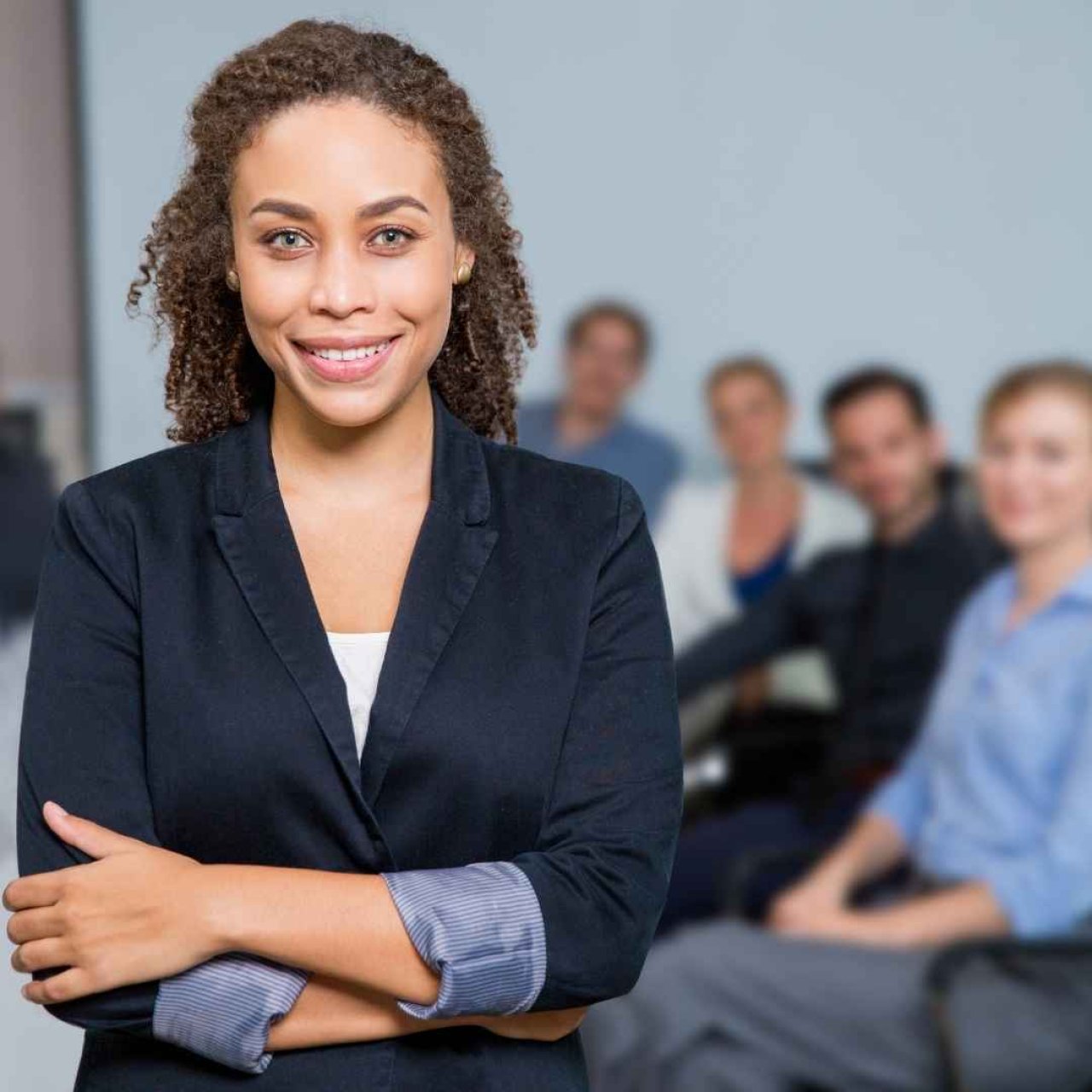 Woman in a suit and folded arms smiling into the camera, with a room full of colleagues blurred behind her smiling at the camera.