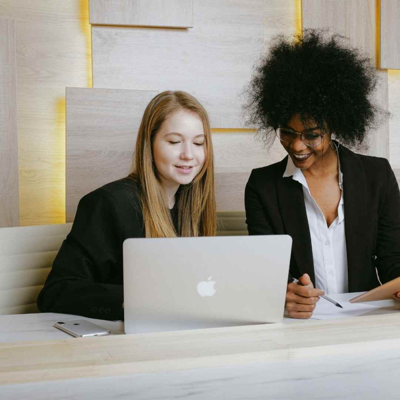 The five workplace gremlins - Two women looking at a laptop with a cool wooden light effect behind them.